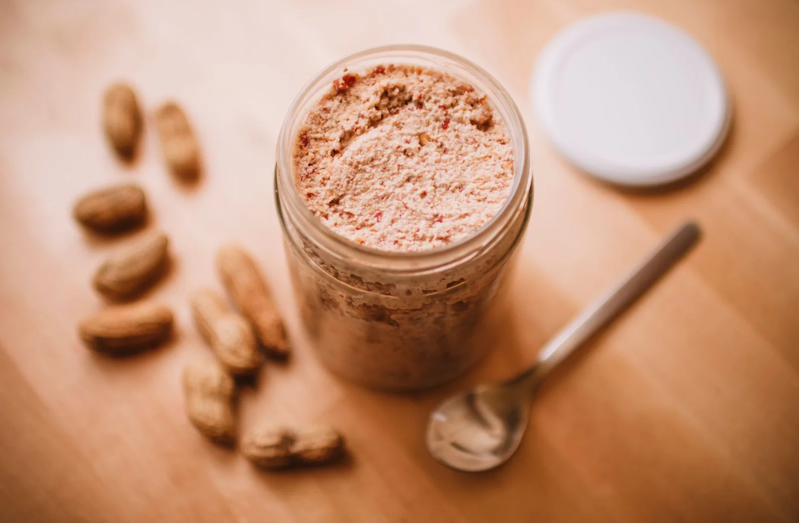 An open jar of peanut butter beside a spoon and scattered peanuts on a wooden table